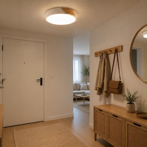 May include: A well-lit entryway with a light-colored rug and wooden furniture. A round mirror and a ceiling light with a wood accent are visible. A coat rack with coats and a purse hangs on the wall. A vase with greenery sits on a wooden cabinet.