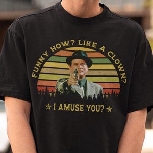 Puede incluir: Camiseta negra con un gráfico retro de un hombre con una pistola, sobre un diseño de puesta de sol. El texto dice "FUNNY HOW? LIKE A CLOWN?" y "I AMUSE YOU?" Una camiseta casual y elegante.