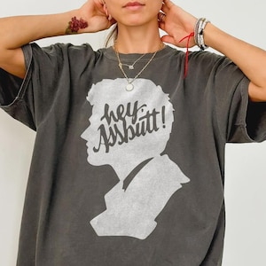 Könnte beinhalten: Dunkelgraues T-Shirt mit einer weißen Silhouette eines Personenprofils und dem Text "hey Assbutt!". Der Text ist handschriftlich. Das T-Shirt hat kurze Ärmel und eine lockere Passform.
