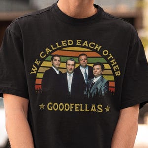 Puede incluir: Camiseta negra con un diseño de cuatro hombres con trajes sobre un fondo de puesta de sol, con el texto "WE CALLED EACH OTHER" y "GOODFELLAS". La camiseta es de cuello redondo.