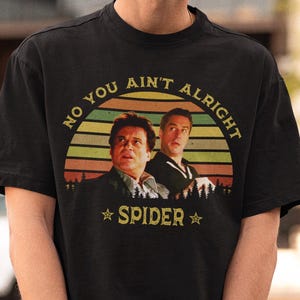Puede incluir: Camiseta negra con un gráfico retro. El diseño presenta el texto "NO YOU AIN'T ALRIGHT" y "SPIDER", junto con una imagen de dos hombres. El fondo es una puesta de sol. Una camiseta clásica.