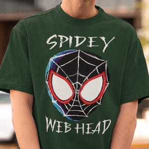 May include: Dark green t-shirt featuring a Spiderman-inspired graphic. The design includes a spider web and the words "Spidey Web Head" in white. The shirt is worn by a person wearing a black cap and sunglasses.