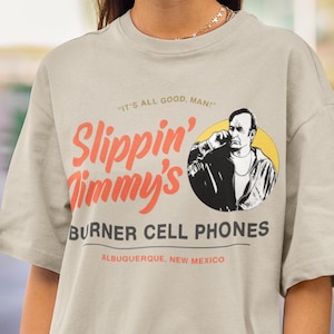 Peut inclure: T-shirt beige avec l'inscription "Slippin' Jimmy's BURNER CELL PHONES" en rouge et noir, avec un dessin d'homme sur un cercle jaune. Le texte "IT'S ALL GOOD, MAN!" est au-dessus du logo, et "ALBUQUERQUE, NEW MEXICO" est en dessous.