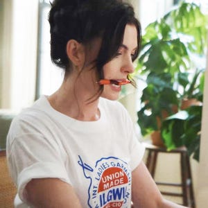 May include: White t-shirt with the text "INT. LADIES GARMENT WORKERS UNION MADE ILGWU AFL-CIO" in blue and red. An orange pen is held in the mouth. The person is in a room with a plant in the background.