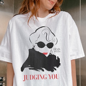 Può includere: T-shirt bianco con una grafica di una donna con occhiali da sole e la scritta "JUDGING YOU" in rosso. La maglietta presenta anche la scritta "The Devil Wears Prada".