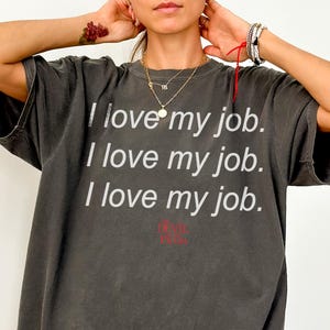 May include: A dark grey Comfort Colors t-shirt with the text "I love my job" repeated three times in white. The shirt also has the text "hell yeah" in red. The shirt is a Pepper colour.