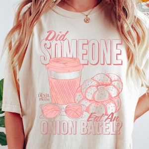 May include: Ivory Comfort Colors tee with a pink graphic. The graphic includes a coffee cup, sunglasses, and a pile of bagels. Text on the shirt reads "Did Someone Eat An Onion Bagel?" and "The Devil Prada."