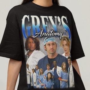 May include: Black t-shirt featuring a cast photo from the television show Grey's Anatomy. The shirt has the show's title in a blue and white design across the top. The cast photo includes several actors in scrubs and lab coats.