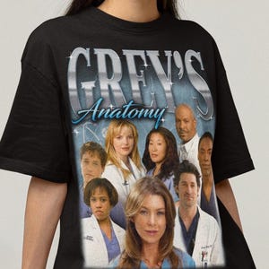 May include: Black t-shirt featuring a graphic of the cast from the television show "Grey's Anatomy." The show's title is displayed in large, silver lettering above the cast image. The shirt is worn by a person.