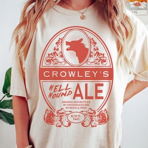 Puede incluir: Una camiseta color crema con un gran gráfico rojo de un sabueso del infierno aullando dentro de un marco ovalado. El texto dice CROWLEY'S HELL HOUND ALE, BREWED AND BOTTLED BY CROSSROADS INN ROSEDALE, MISS, y EST. 1661, rodeado de adornos.