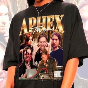 May include: A black short-sleeve t-shirt featuring a graphic print of Aphex Twin. The design includes the text APHEX TWIN in gold lettering above a collage of multiple photographs of the artist, including one where they are holding a mask.