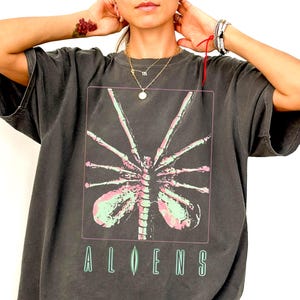 May include: A charcoal grey short-sleeve t-shirt featuring a rectangular graphic of a pale green and pink skeletal creature with long limbs. Below the graphic, the word ALIENS is printed in a stylised green font with a vertical line accent.