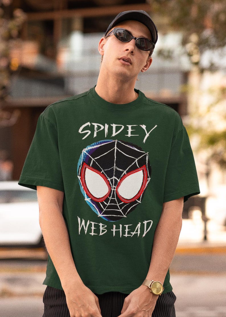 May include: Dark green t-shirt featuring a Spiderman-inspired graphic. The design includes a spider web and the words "Spidey Web Head" in white. The shirt is worn by a person wearing a black cap and sunglasses.