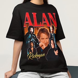 May include: A black short-sleeve t-shirt featuring a graphic print of actor Alan Rickman in various roles. The name ALAN is printed in large red letters at the top, and the name Rickman is printed in a gold cursive script at the bottom of the graphic.