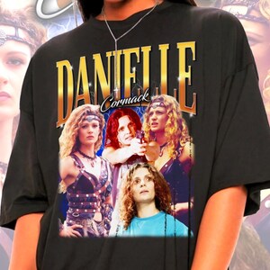 Peut inclure: Un t-shirt noir à manches courtes avec une impression graphique de l'actrice Danielle Cormack. Le motif comprend le nom DANIELLE CORMACK en grandes lettres dorées au-dessus de plusieurs images de l'actrice aux cheveux bouclés.