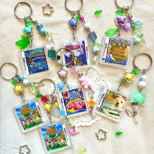Può includere: Portachiavi con custodie di giochi in miniatura, tra cui "Pokemon Mystery Dungeon," "Zelda," "Animal Crossing," e "Kirby." Ogni portachiavi ha un anello argentato, perline colorate e ciondoli decorativi.
