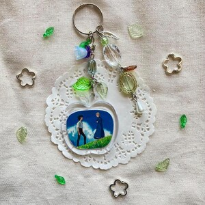 May include: A clear apple-shaped keychain with a scene of two figures on a hill. The keychain has a silver ring and is adorned with beads in various colors, including green, blue, and clear. The keychain is on a white doily.
