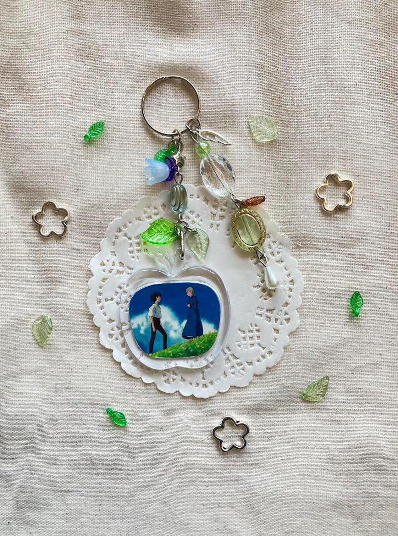May include: A clear apple-shaped keychain with a scene of two figures on a hill. The keychain has a silver ring and is adorned with beads in various colors, including green, blue, and clear. The keychain is on a white doily.