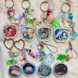May include: A collection of anime-themed keychains. The keychains feature various characters from Studio Ghibli films, with colorful beads and charms. The keychains are in the shape of apples and circles, with silver key rings.