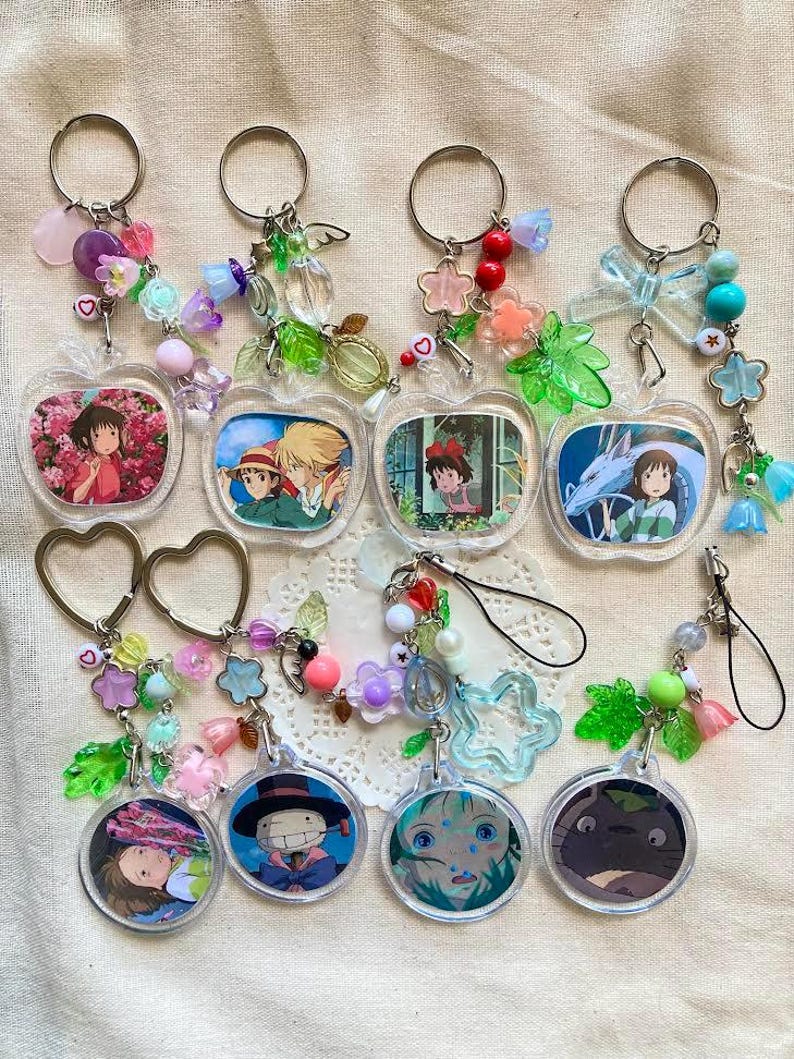 May include: A collection of anime-themed keychains. The keychains feature various characters from Studio Ghibli films, with colorful beads and charms. The keychains are in the shape of apples and circles, with silver key rings.