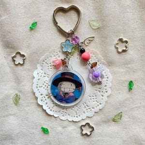 May include: A keychain featuring a circular charm with a cartoon character in a black hat and blue outfit. The keychain has a heart-shaped ring, decorative beads, and a small wing charm. The charm is attached to a white lace doily.