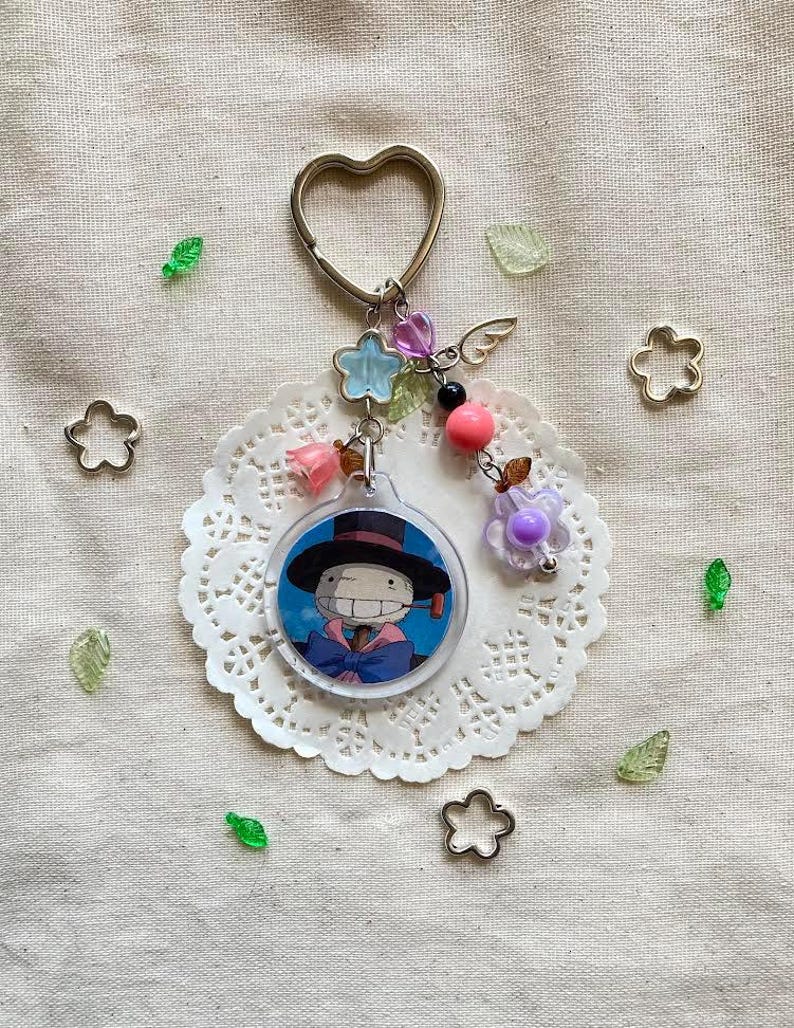 May include: A keychain featuring a circular charm with a cartoon character in a black hat and blue outfit. The keychain has a heart-shaped ring, decorative beads, and a small wing charm. The charm is attached to a white lace doily.