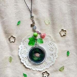 May include: A clear, round charm with a forest scene and a small animal inside, attached to a black cord. The charm is adorned with green leaves, pink roses, and beads. Decorative metal flower shapes and leaf accents are scattered around.