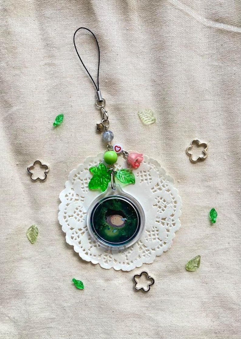 May include: A clear, round charm with a forest scene and a small animal inside, attached to a black cord. The charm is adorned with green leaves, pink roses, and beads. Decorative metal flower shapes and leaf accents are scattered around.