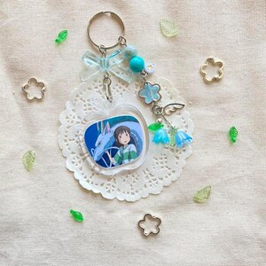 May include: A clear acrylic keychain featuring a cartoon image of a girl and a dragon. The keychain is attached to a silver keyring with a light blue bow, beads, and flower charms. The keychain is on a white doily with decorative accents.