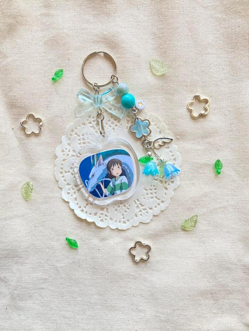 May include: A clear acrylic keychain featuring a cartoon image of a girl and a dragon. The keychain is attached to a silver keyring with a light blue bow, beads, and flower charms. The keychain is on a white doily with decorative accents.