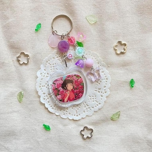 May include: A clear apple-shaped keychain with a picture of a girl in a pink dress surrounded by pink flowers. The keychain has a silver ring and is adorned with pink, purple, and clear beads, a small heart charm, and a butterfly charm. The keychain rests on a white doily.
