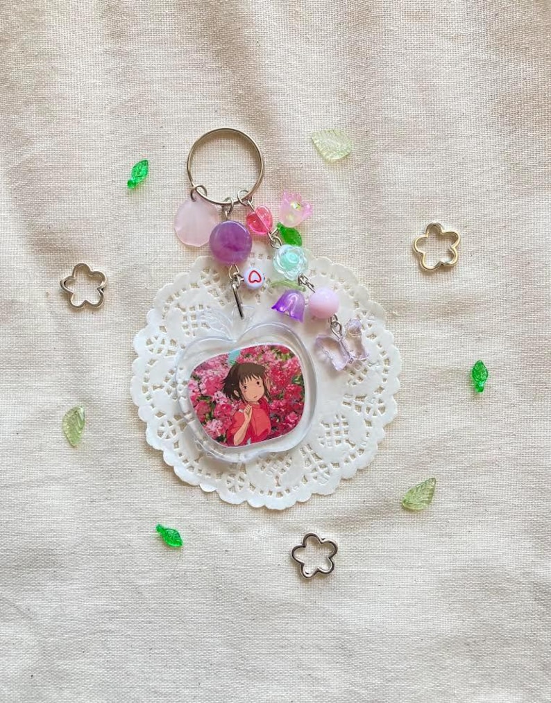 May include: A clear apple-shaped keychain with a picture of a girl in a pink dress surrounded by pink flowers. The keychain has a silver ring and is adorned with pink, purple, and clear beads, a small heart charm, and a butterfly charm. The keychain rests on a white doily.
