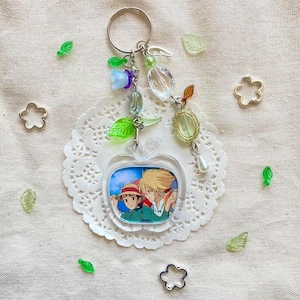 May include: A clear acrylic apple-shaped keychain featuring a colorful anime illustration of two characters. The keychain is adorned with various charms, including green leaves, clear beads, and a silver key. The keychain is on a white doily with scattered decorative elements.