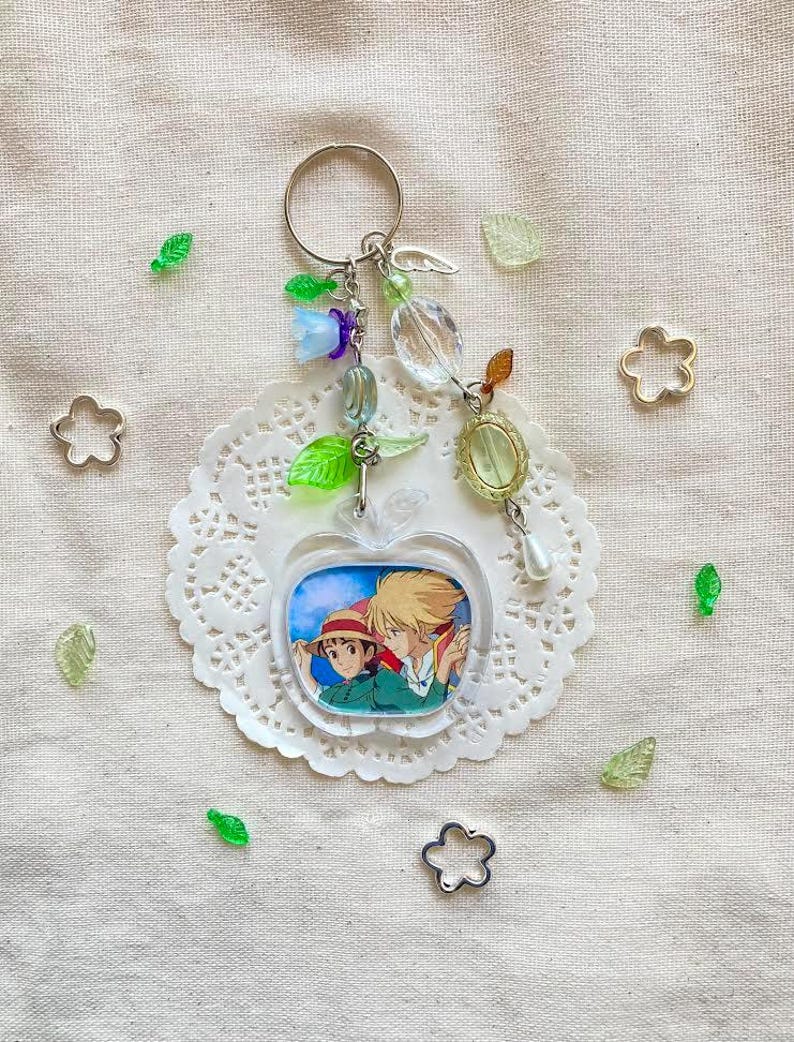 May include: A clear acrylic apple-shaped keychain featuring a colorful anime illustration of two characters. The keychain is adorned with various charms, including green leaves, clear beads, and a silver key. The keychain is on a white doily with scattered decorative elements.