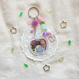 May include: A clear acrylic keychain with a floral design inside an apple shape. The keychain has a silver ring and is adorned with pink, purple, and clear beads, along with a small heart charm. Decorative elements include flower and leaf-shaped embellishments.