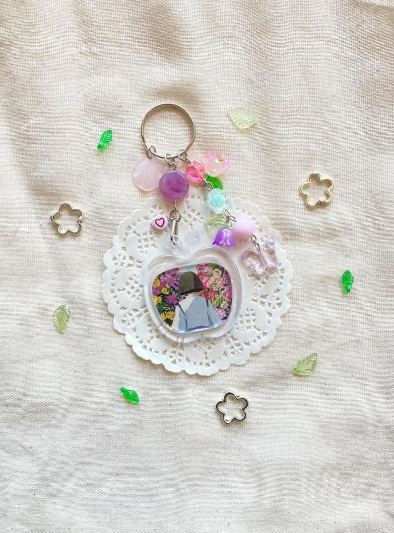 May include: A clear acrylic keychain with a floral design inside an apple shape. The keychain has a silver ring and is adorned with pink, purple, and clear beads, along with a small heart charm. Decorative elements include flower and leaf-shaped embellishments.