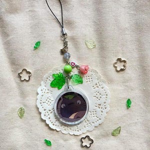 May include: A clear acrylic charm featuring a cartoon character with a dark purple body and large eyes. The charm is attached to a black lanyard with decorative beads, a pink rose, and green leaf accents. The charm rests on a white lace doily.
