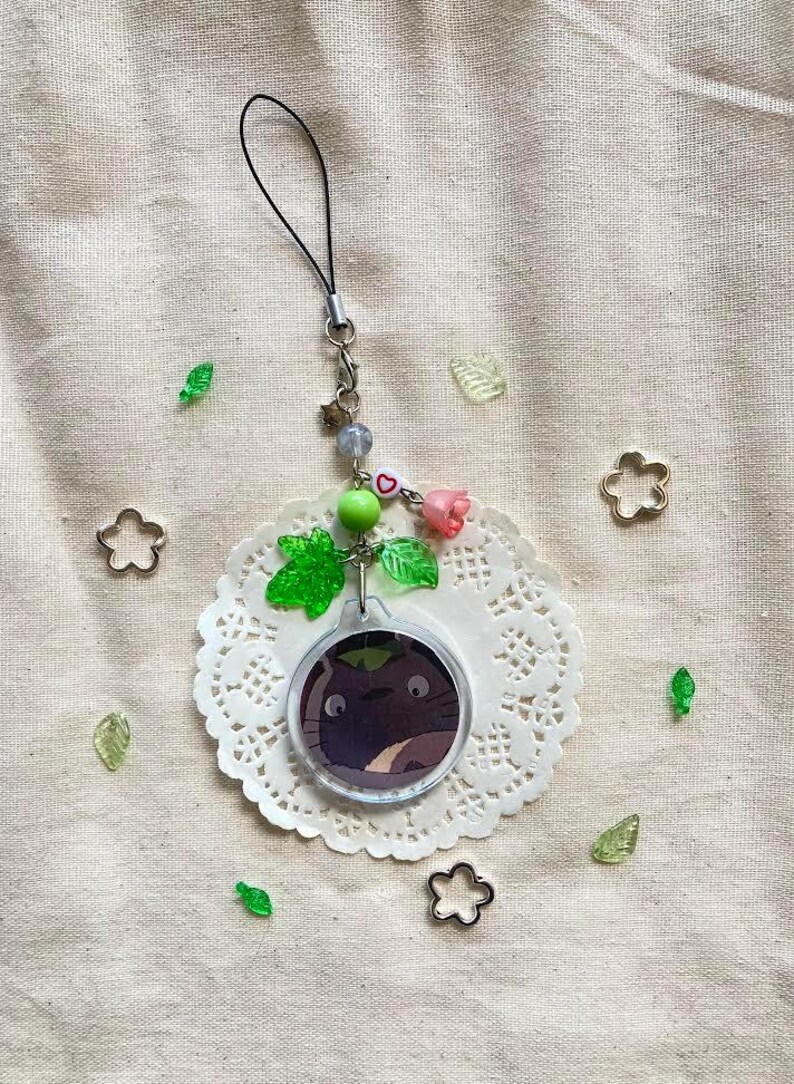 May include: A clear acrylic charm featuring a cartoon character with a dark purple body and large eyes. The charm is attached to a black lanyard with decorative beads, a pink rose, and green leaf accents. The charm rests on a white lace doily.