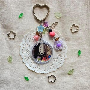 May include: A keychain featuring a circular charm with a character from Spirited Away, surrounded by decorative beads, flowers, and a heart-shaped keyring. The charm is set on a white lace doily, with scattered floral and leaf accents.