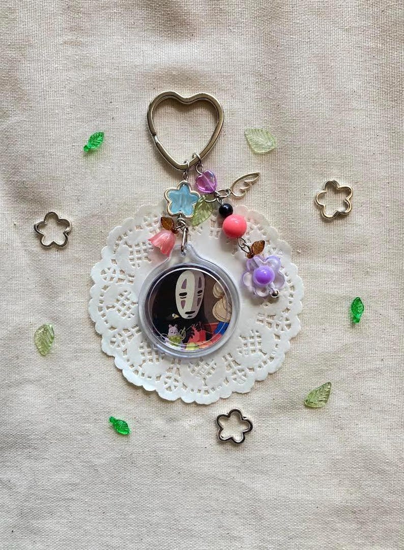 May include: A keychain featuring a circular charm with a character from Spirited Away, surrounded by decorative beads, flowers, and a heart-shaped keyring. The charm is set on a white lace doily, with scattered floral and leaf accents.