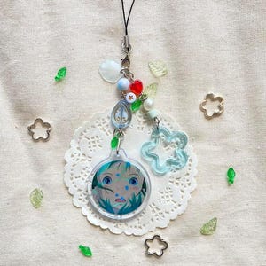 May include: A handmade charm with a cartoon face, blue and white beads, and a clear blue flower. The charm is attached to a black cord. Decorative elements include small metal flower shapes and green leaf-shaped beads.