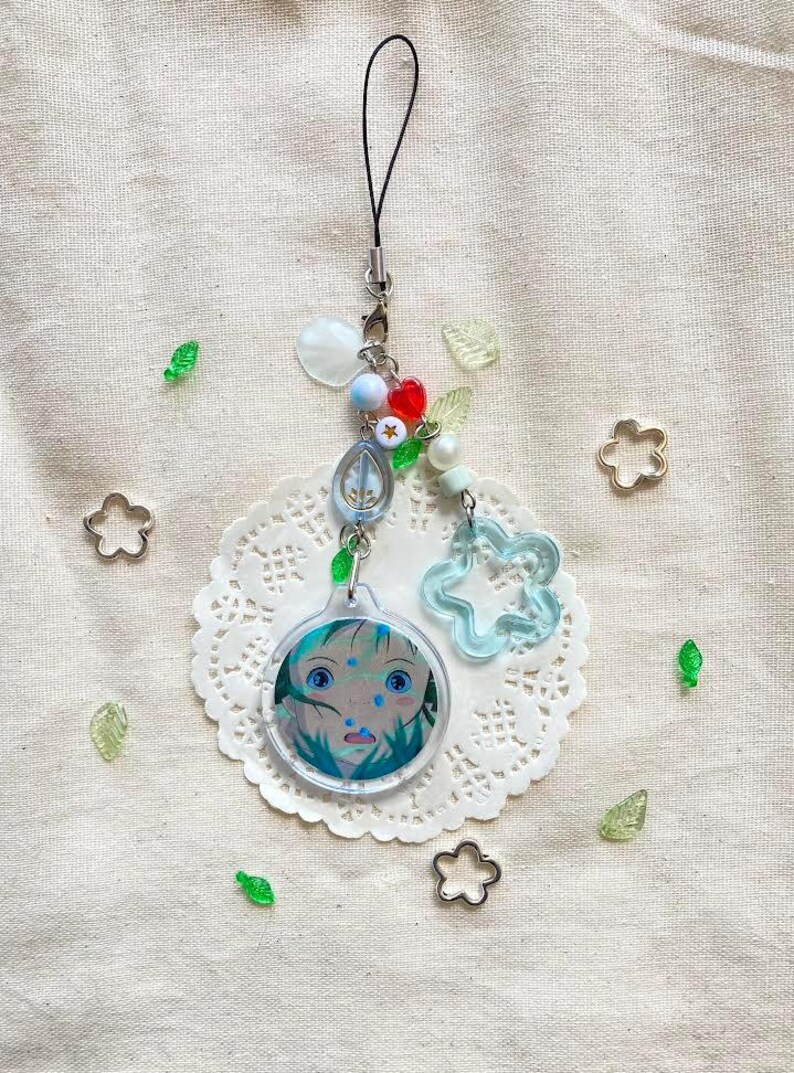 May include: A handmade charm with a cartoon face, blue and white beads, and a clear blue flower. The charm is attached to a black cord. Decorative elements include small metal flower shapes and green leaf-shaped beads.