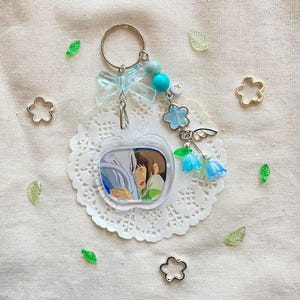 May include: A clear apple-shaped keychain featuring a cartoon image of two characters. The keychain has a silver ring, blue beads, a blue bow, and flower and bell charms. Decorative elements include small flower and leaf-shaped embellishments.