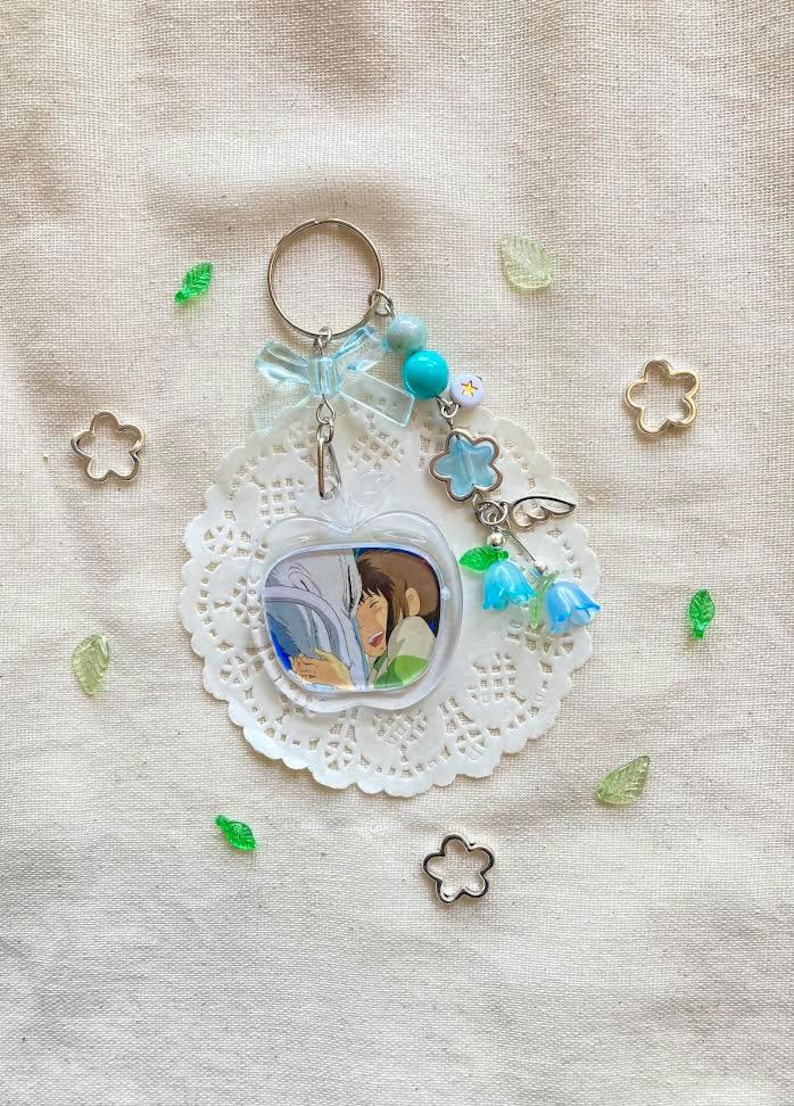 May include: A clear apple-shaped keychain featuring a cartoon image of two characters. The keychain has a silver ring, blue beads, a blue bow, and flower and bell charms. Decorative elements include small flower and leaf-shaped embellishments.