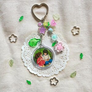 May include: A keychain with a heart-shaped ring and a charm featuring an anime-style illustration of two people. The keychain is adorned with colorful beads, flowers, and green leaf-shaped accents. The charm is round and clear, with a white lace doily underneath.