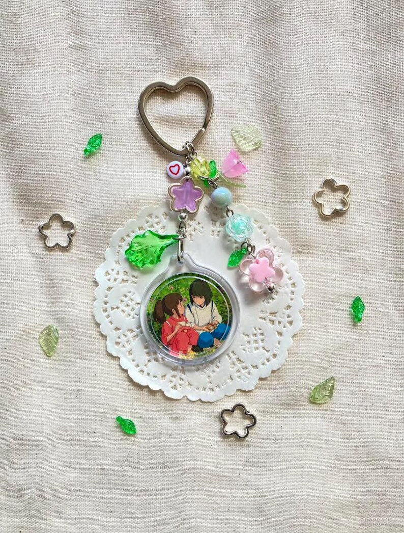 May include: A keychain with a heart-shaped ring and a charm featuring an anime-style illustration of two people. The keychain is adorned with colorful beads, flowers, and green leaf-shaped accents. The charm is round and clear, with a white lace doily underneath.