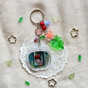 May include: A clear acrylic keychain featuring a cartoon image of a girl in a red bow. The keychain is apple-shaped and adorned with pink and clear flower charms, red beads, and a silver keyring. Decorative elements include green leaf and flower-shaped accents.