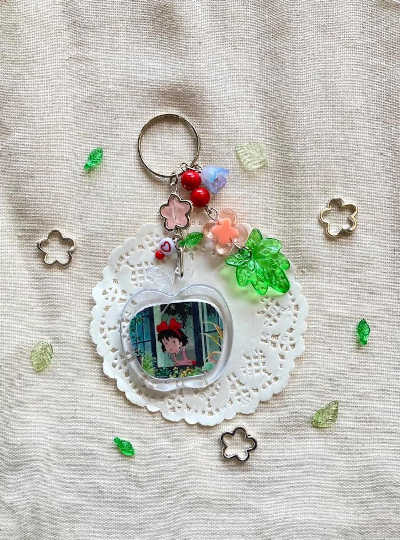 May include: A clear acrylic keychain featuring a cartoon image of a girl in a red bow. The keychain is apple-shaped and adorned with pink and clear flower charms, red beads, and a silver keyring. Decorative elements include green leaf and flower-shaped accents.