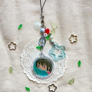 May include: A clear acrylic charm with a cartoon image of a girl, attached to a beaded charm with a black cord. The charm features blue, red, and white beads, a star, and a clear blue flower. Decorative green leaf and flower shapes are scattered around.