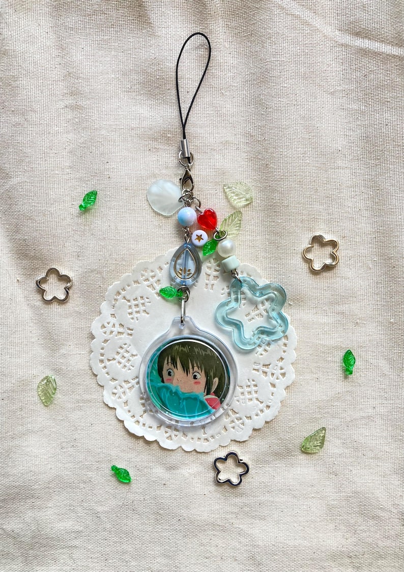 May include: A clear acrylic charm with a cartoon image of a girl, attached to a beaded charm with a black cord. The charm features blue, red, and white beads, a star, and a clear blue flower. Decorative green leaf and flower shapes are scattered around.